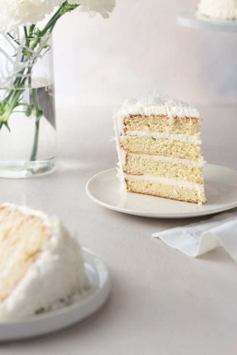 Straight on view of a slice of coconut layer cake with Swiss meringue buttercream on a plate surrounded by another slice of cake, plates, a napkin, a vase of white carnation flowers and the cake on a cake stand with a cream colored surface and light textured background.