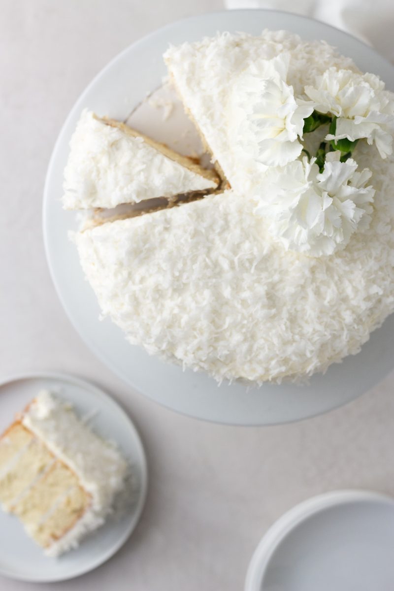 Overhead view of a sliced coconut layer cake with Swiss meringue buttercream on a cake stand topped with white carnation flowers with a cream colored surface and light textured background.
