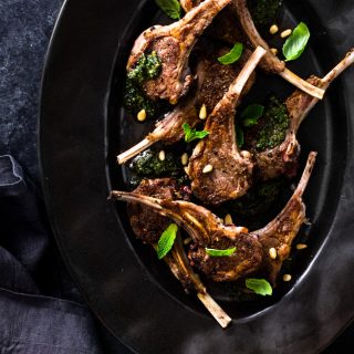 Overhead shot of a black serving platter topped with seared baby lamb chops and mint pesto on a dark, textured surface.