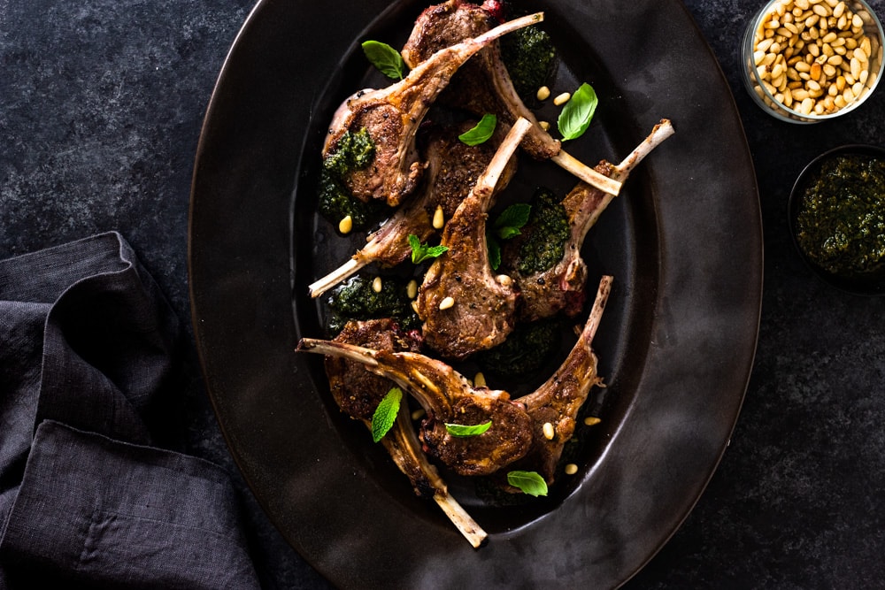 Overhead shot of a black serving dish with seared baby lamb chops, mint pesto, pine nuts and fresh herbs on a dark textured background.