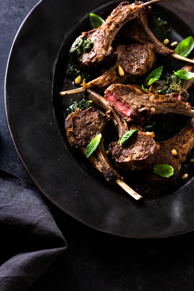 Overhead shot of a black serving dish with seared baby lamb chops (one with piece sliced open), mint pesto, pine nuts and fresh herbs on a dark textured background.