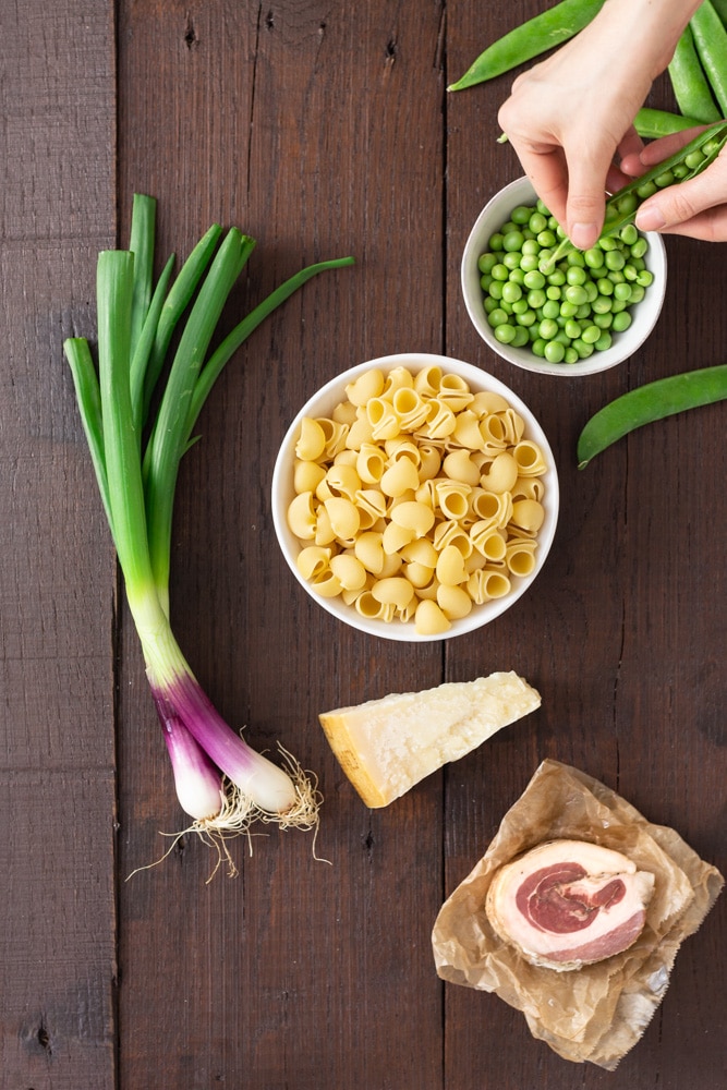 Overhead view of prep for pasta with peas and pancetta, including shell pasta, spring onions, pancetta, parmesan cheese and fresh peas on a dark wood surface.