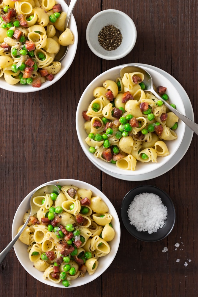  Overhead view of three bowls of shell pasta with peas and pancetta with spoons and small bowls of salt and pepper on a dark wood surface.