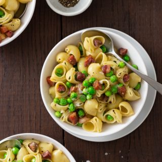 Overhead view of three bowls of shell pasta with peas and pancetta with spoons and small bowls of salt and pepper on a dark wood surface.