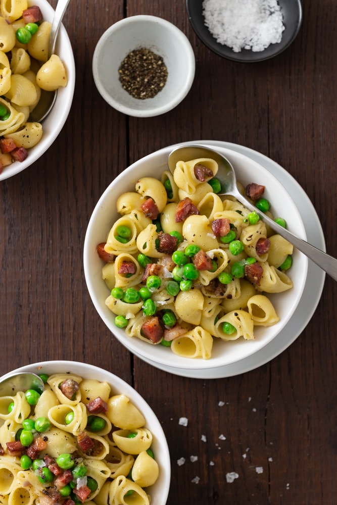 Overhead view of three bowls of shell pasta with peas and pancetta with spoons and small bowls of salt and pepper on a dark wood surface.