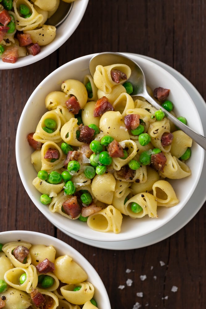  Overhead view of three bowls of shell pasta with peas and pancetta with spoons and small bowls of salt and pepper on a dark wood surface.