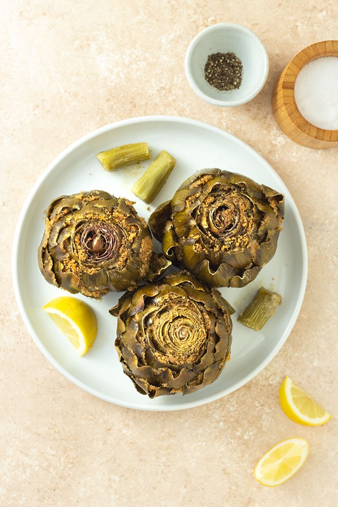 Overhead view of Italian Stuffed Artichokes filled with breadcrumbs, cheese, garlic and parsley on a white plate surrounded by lemon wedges, and bowls of salt and pepper on a beige textured surface.