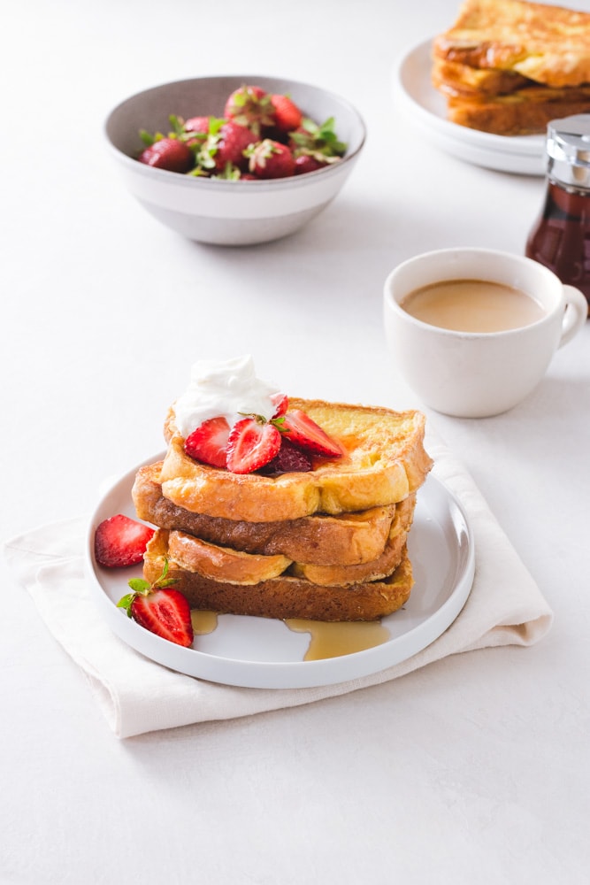 ¾ angled view of a stack of Brioche French Toast topped with macerated strawberries and whipped mascarpone cream on a white plate on a napkin on a white surface with strawberries, a cup of coffee, maple syrup and a plate of more French toast in the background.