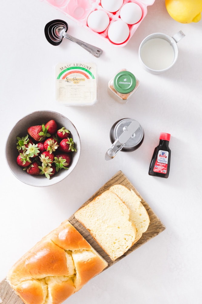 Overhead view of ingredients for French toast, including eggs, a lemon, cinnamon, maple syrup, vanilla extract, brioche bread, strawberries and mascarpone cream.