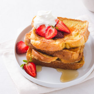3/4 angled view of a stack of Brioche French toast topped with macerated strawberries and whipped mascarpone cream on a white plate on a napkin on a white background next to a cup of coffee.