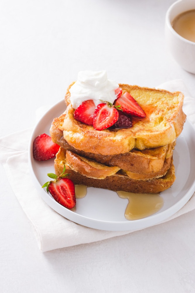 ¾ angled view of a stack of Brioche French toast topped with macerated strawberries and whipped mascarpone cream on a white plate on a napkin on a white background next to a cup of coffee.