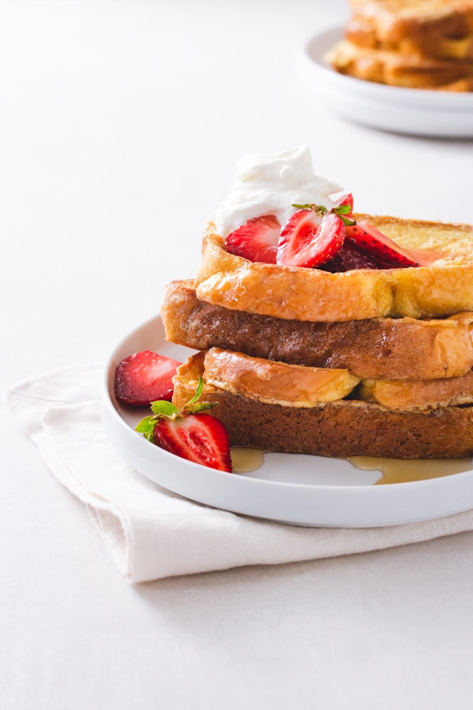 Straight on view of a stack of Brioche French Toast topped with macerated strawberries and whipped mascarpone cream on a white plate on a napkin on a white surface with a plate of more French toast in the background.