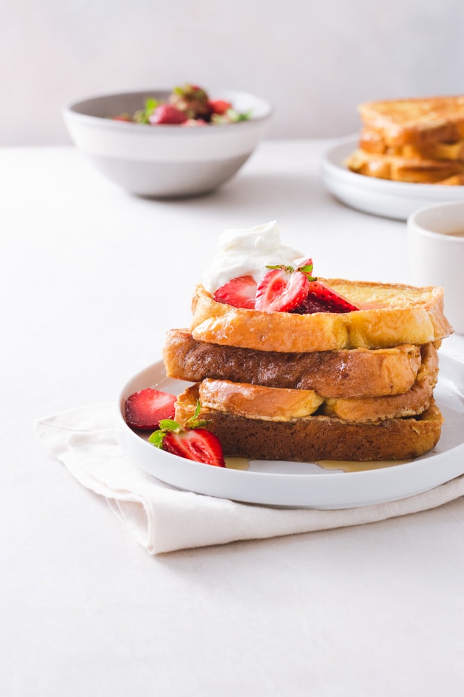 Straight on view of a stack of Brioche French Toast topped with macerated strawberries and whipped mascarpone cream on a white plate on a napkin on a white surface with strawberries, a cup of coffee, and a plate of more French toast in the background.