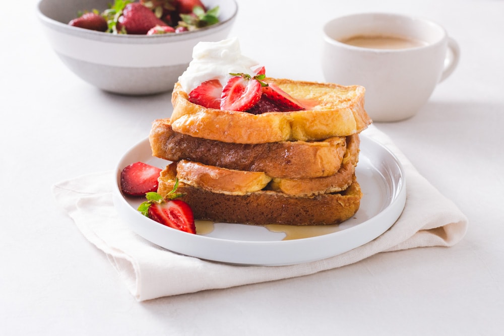 Straight on view of a stack of Brioche French Toast topped with macerated strawberries and whipped mascarpone cream on a white plate on a napkin on a white surface with a cup of coffee and bowl of strawberries in the background.