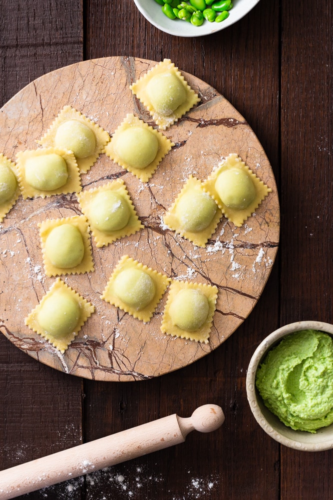 Overhead image of raw fava bean and ricotta ravioli on a light brown marble round on a dark wood surface surrounded by a bowl of filling, a bowl of fava beans and a small wooden rolling pin.