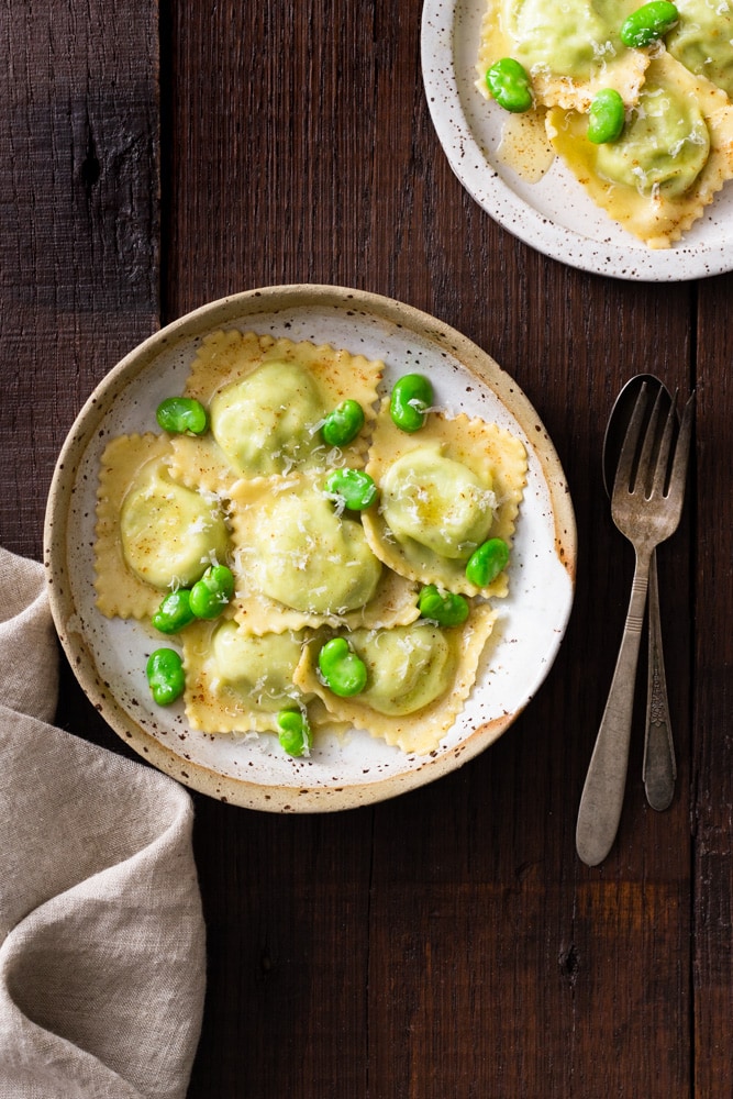 Overhead image of a rustic ceramic bowl with fava bean and ricotta ravioli in brown butter sauce with whole fava beans and parmesan cheese on top, surrounded by rustic silverware (fork and spoon), a beige dish towel and another plate of pasta on a dark wood surface.