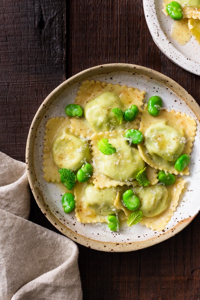 Overhead image of a rustic ceramic bowl with fava bean and ricotta ravioli in brown butter sauce with whole fava beans, mint and parmesan cheese on top, surrounded by a beige dish towel and another plate of pasta on a dark wood surface.