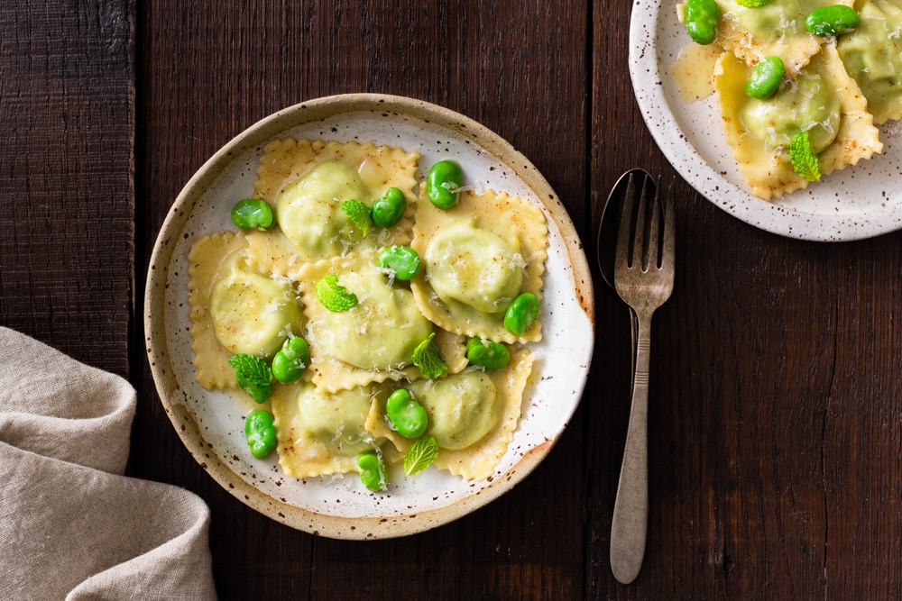 Overhead image of a rustic ceramic bowl with fava bean and ricotta ravioli in brown butter sauce with whole fava beans, mint and parmesan cheese on top, surrounded by rustic silverware (fork and spoon), a beige dish towel and another plate of pasta on a dark wood surface.