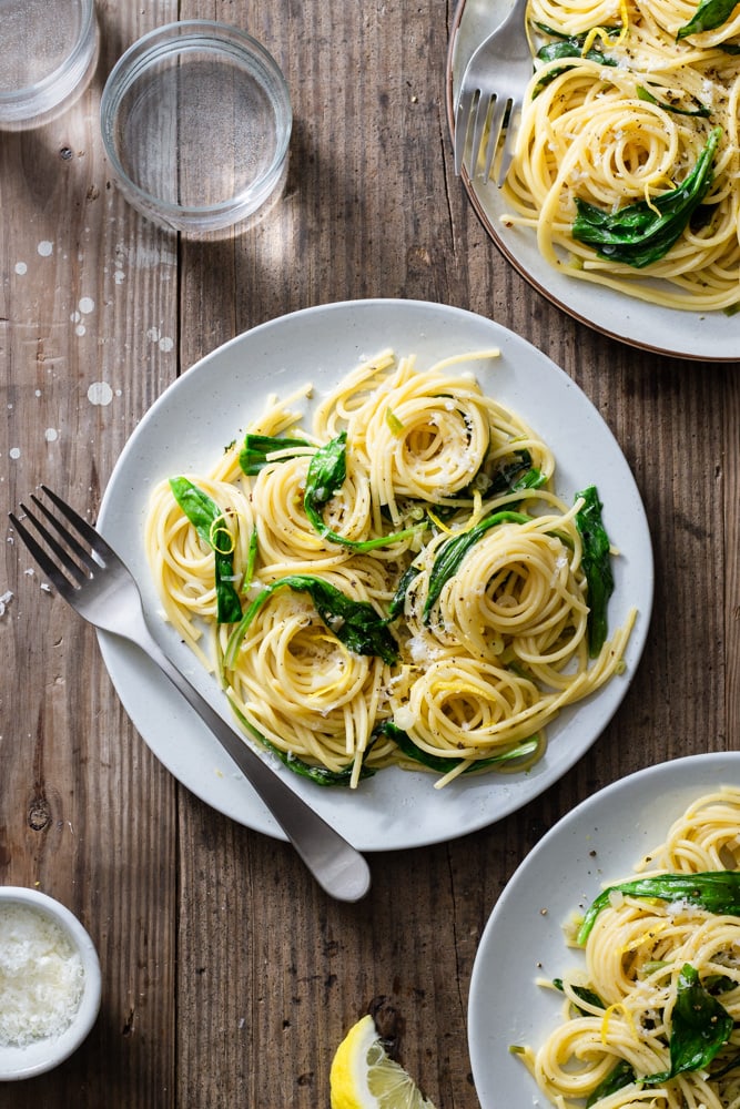 Overhead view of three plates of spaghetti pasta with ramps surrounded by forks, glasses, grated parmesan cheese and lemon wedges on a rustic wood surface.