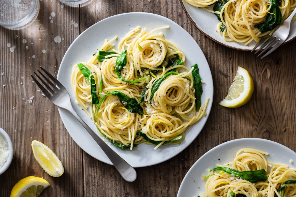 Overhead view of three plates of spaghetti pasta with ramps surrounded by forks, glasses, grated parmesan cheese and lemon wedges on a rustic wood surface. 	