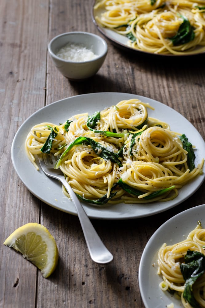 ¾ angled view of three plates of spaghetti pasta with ramps surrounded by forks, glasses, grated parmesan cheese and lemon wedges on a rustic wood surface. 	