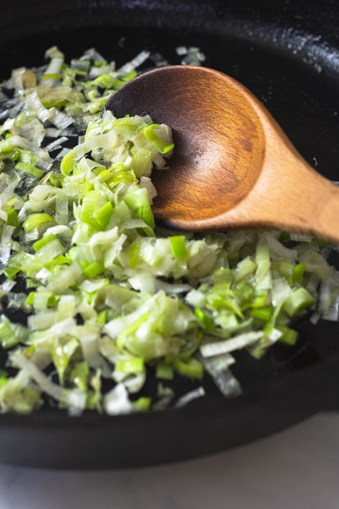 Angled close-up view of sliced leeks sauteing in a cast iron pan with a wooden spoon.