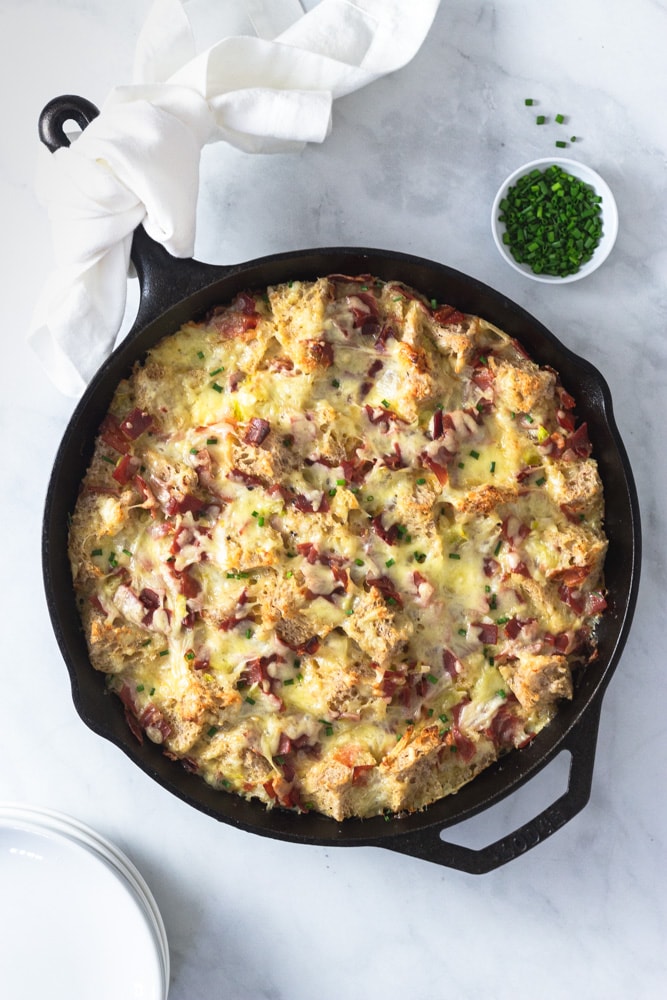 Overhead shot of a Ham and Cheese Strata made with Bayonne Ham in a cast iron pan on a marble surface surrounded by a bowl of chopped chives and a stack of white plates.