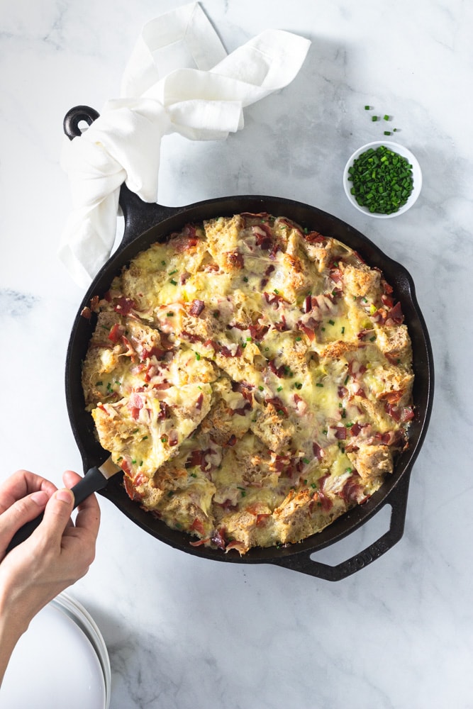 Overhead shot of a slice being lifted out of a Ham and Cheese Strata made with Bayonne Ham in a cast iron pan on a marble surface surrounded by a bowl of chopped chives and a stack of white plates.