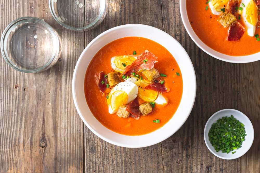 Overhead shot of a Spanish chilled tomato soup, Salmorejo topped with croutons, hard boiled eggs, crispy ham, and chives on a rustic grey wood surface surrounded by multiple bowls of soup, water glasses and a bowl of cut chives.
