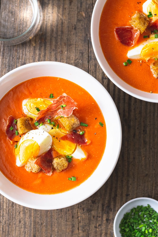 Overhead shot of a Spanish chilled tomato soup, Salmorejo topped with croutons, hard boiled eggs, crispy ham, and chives on a rustic grey wood surface surrounded by another bowl of soup, a water glass and a bowl of cut chives.