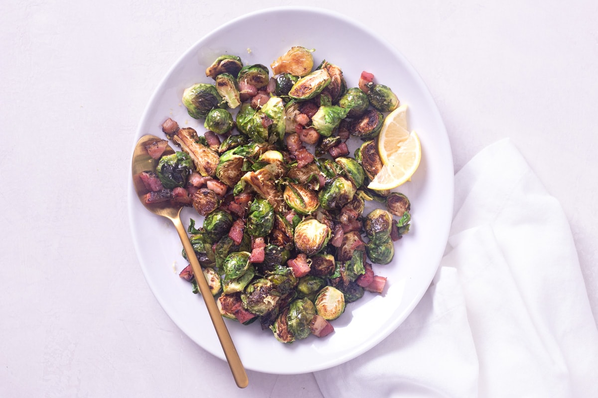 Pan roasted brussels sprouts with pancetta on a white serving platter with a copper spoon on a light surface.