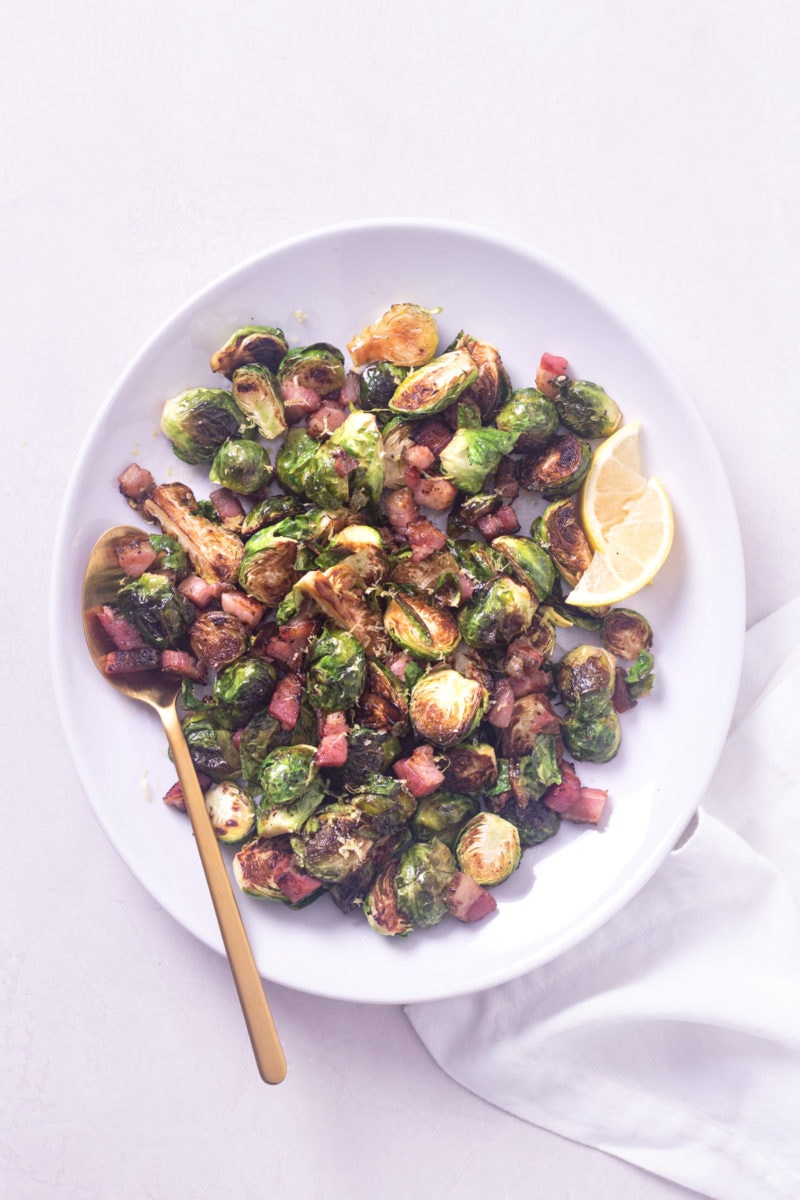 Pan roasted brussels sprouts with pancetta on a white serving platter with a copper spoon on a light surface.