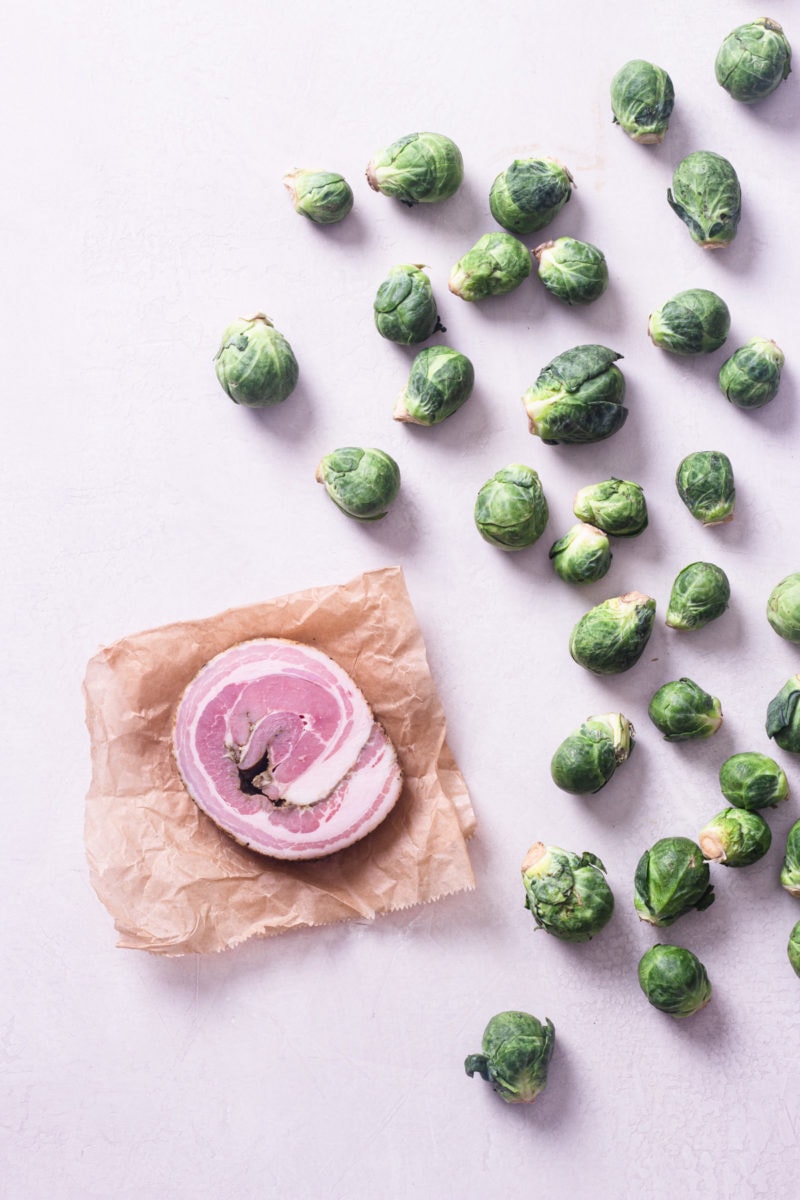 Overhead view of a round of raw pancetta on brown parchment next to whole raw brussels sprouts on a light surface