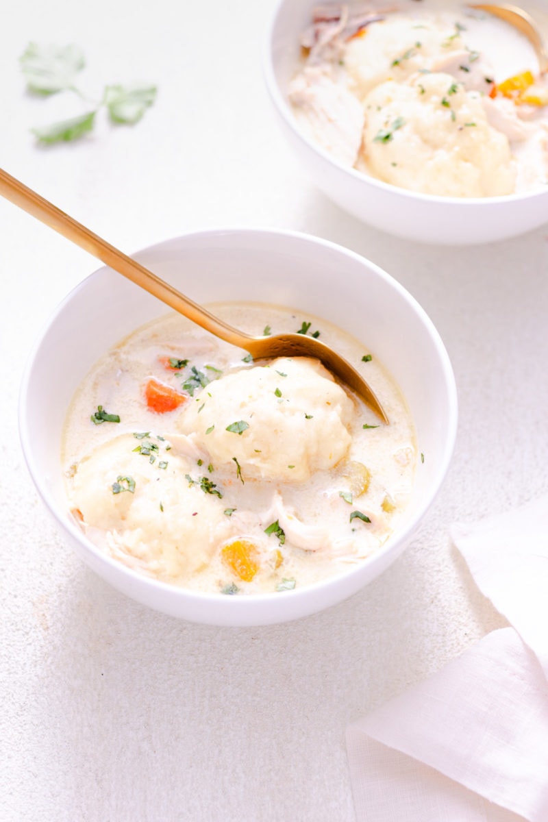 Chicken and Dumpling Soup in a white bowl with a gold spoon, another soup bowl in the background and a white surface.