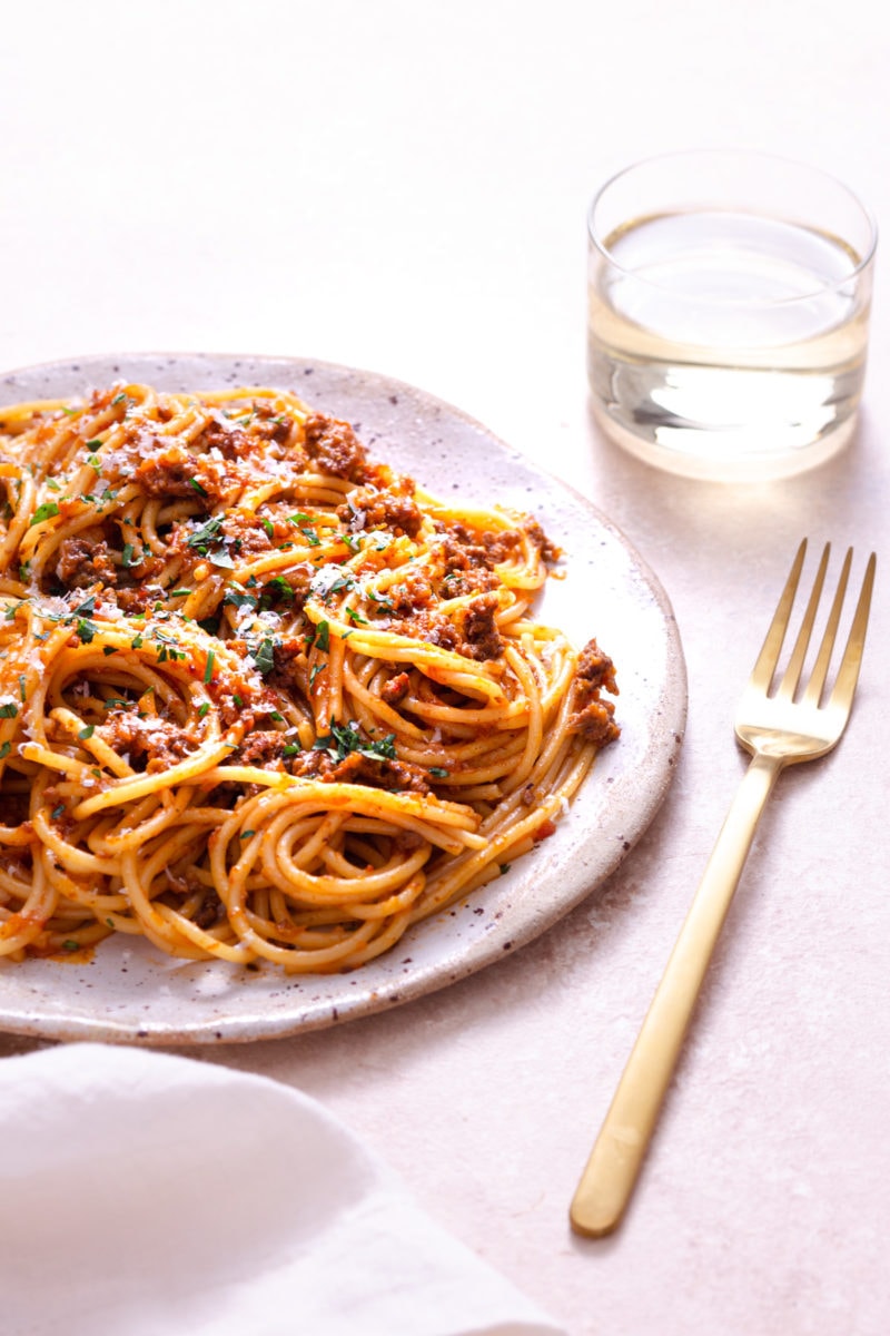 Angled, close up shot of a plate of spaghetti bolognese topped with parsley and parmesan cheese on a light, rustic surface next to a gold fork, cream napkin and glass of white wine.