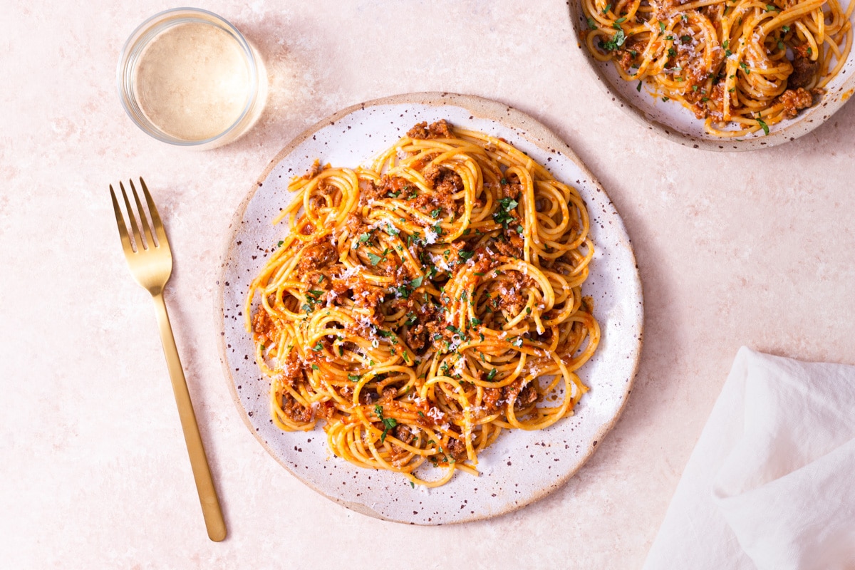 Overhead shot of a plate of spaghetti bolognese topped with parsley and parmesan cheese on a light, rustic surface next to a gold fork, cream napkin and glass of white wine.