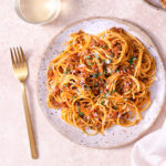 Overhead shot of a plate of spaghetti bolognese topped with parsley and parmesan cheese on a light, rustic surface next to a gold fork, cream napkin and glass of white wine.