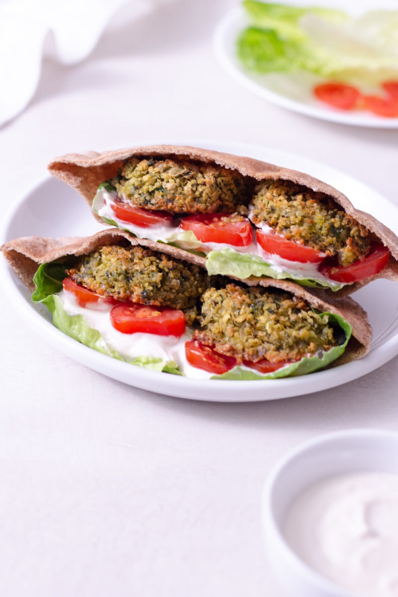 Straight on view of a crispy, baked falafel sandwich with lettuce, tomato and tahini yogurt sauce on a white plate surrounded by the sandwich ingredients and a dish towel on an off white surface.