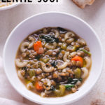 Angled view of Italian Lentil Soup with carrots, celery and spinach, topped with parmesan, and surrounded by another bowl of soup, bread and a dish towel.