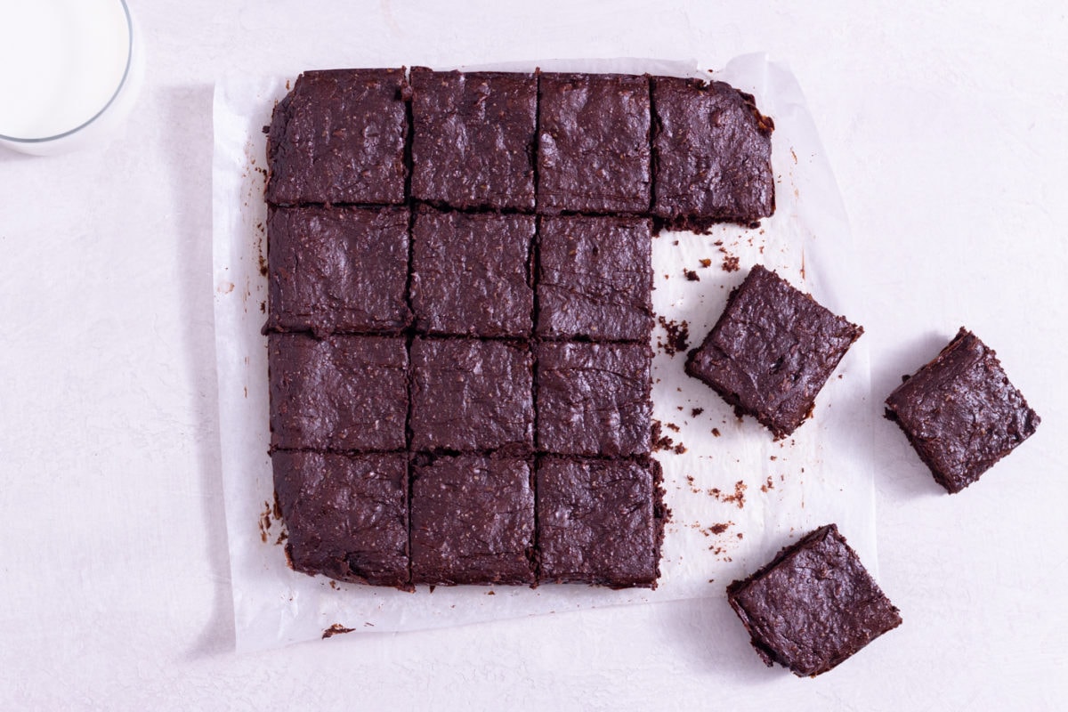 Overhead view of healthy brownies cut into squares on parchment paper next to a glass of milk on a white surface.