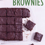 Overhead view of Avocado Brownies cut into squares on parchment paper next to a glass of milk on a white surface.