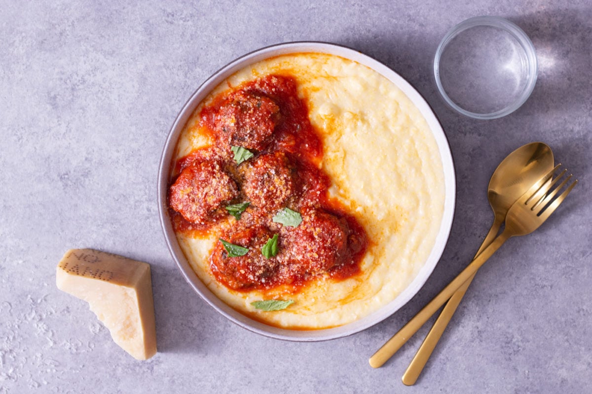 Overhead view of a bowl of polenta topped with meatballs in tomato sauce cooked in pressure cooker next to a glass, gold utensils, and a hunk of parmesan on a light grey surface.