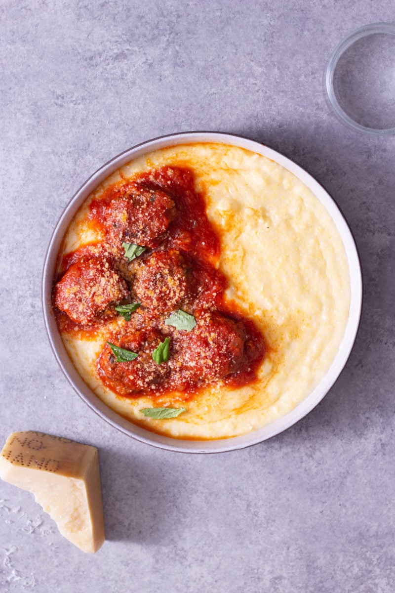 Overhead view of a bowl of polenta topped with meatballs in tomato sauce cooked in pressure cooker next to a glass and a hunk of parmesan on a light grey surface.