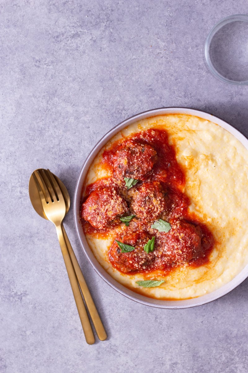 Overhead view of a bowl of polenta topped with Instant Pot Meatballs in tomato sauce next to a glass and gold utensils on a light grey surface.