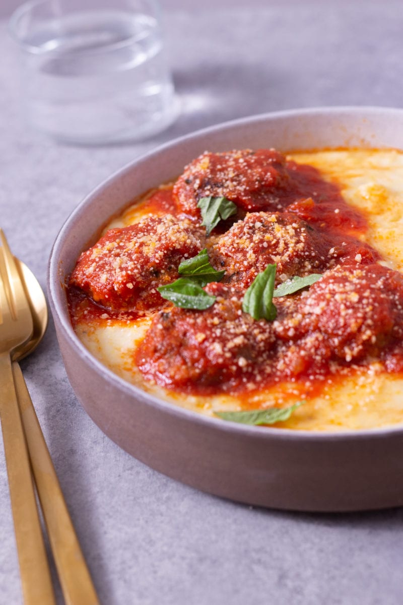 Close up angled view of a bowl of polenta topped with Instant Pot Meatballs in tomato sauce next to gold utensils on a light grey surface.