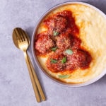 Overhead view of a bowl of polenta topped with Italian meatballs in tomato sauce cooked in a pressure cooker next to gold silverware on a light grey, textured surface.