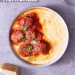 Overhead view of a bowl of polenta topped with Instant Pot Meatballs in tomato sauce next to a glass and a hunk of parmesan on a light grey surface.