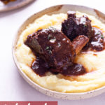 Angled view of a shallow bowl of mashed potatoes topped with Instant Pot Short Ribs with a red wine tomato sauce next to copper utensils on an off white surface.