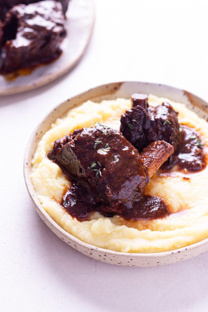 Angled view of a shallow bowl of mashed potatoes topped with Instant Pot Short Ribs with a red wine tomato sauce on an off white surface.