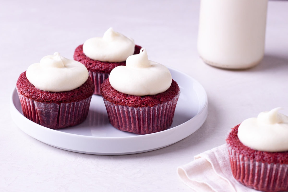 Straight on view of a plate of red velvet cupcakes with cream cheese frosting with a jug of milk in the background.